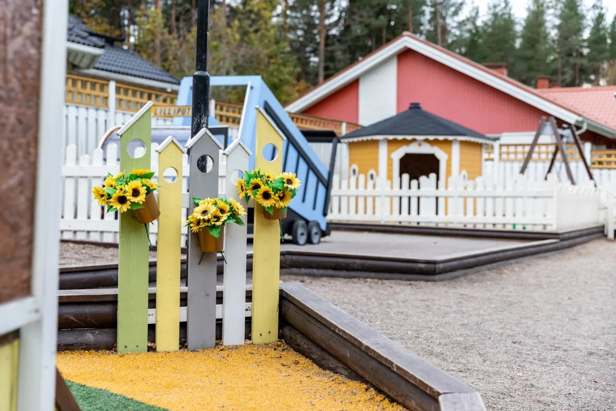 Päiväkoti Lilliputin piha: värikäs aita auringonkukkineen, leikkimökki ja punainen päiväkotirakennus.